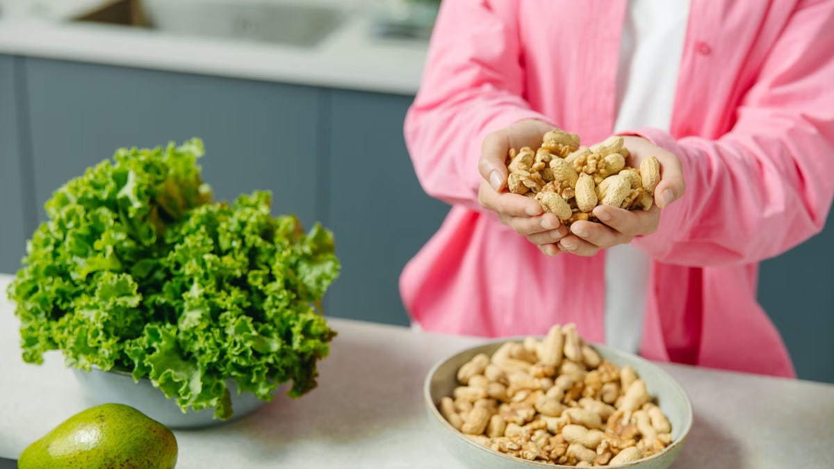 Hands presenting peanuts with a bowl, lettuce, avocado, and tomato nearby on kitchen counter.
